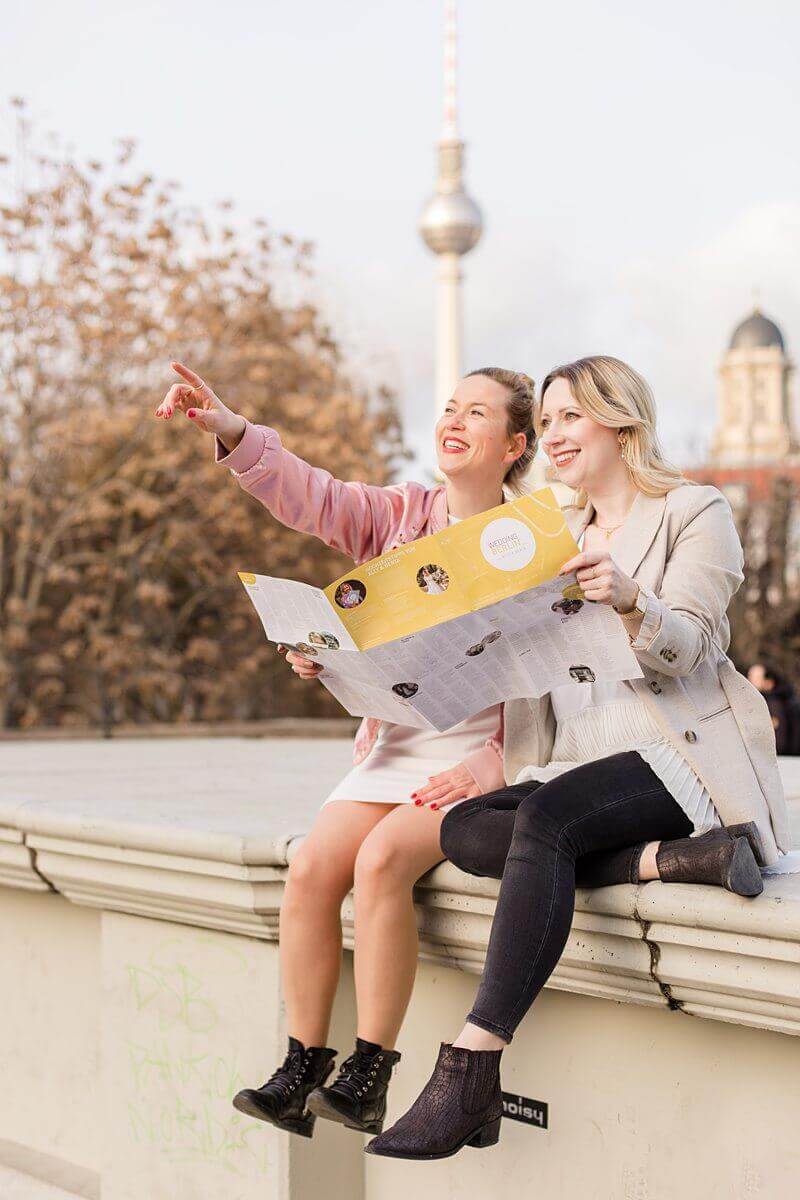 Zwei Frauen sitzen auf einer Mauer vor dem Fernsehturm Berlin, lachen in die Ferne und zeigen auf etwas, dabei halten sie eine Karte fest