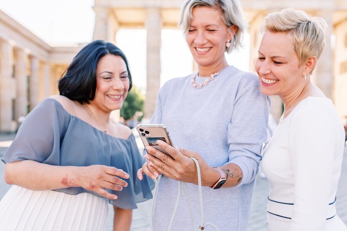 Drei Frauen schauen lachend auf ein Handy, im Hintergrund das Brandenburger Tor Berlin