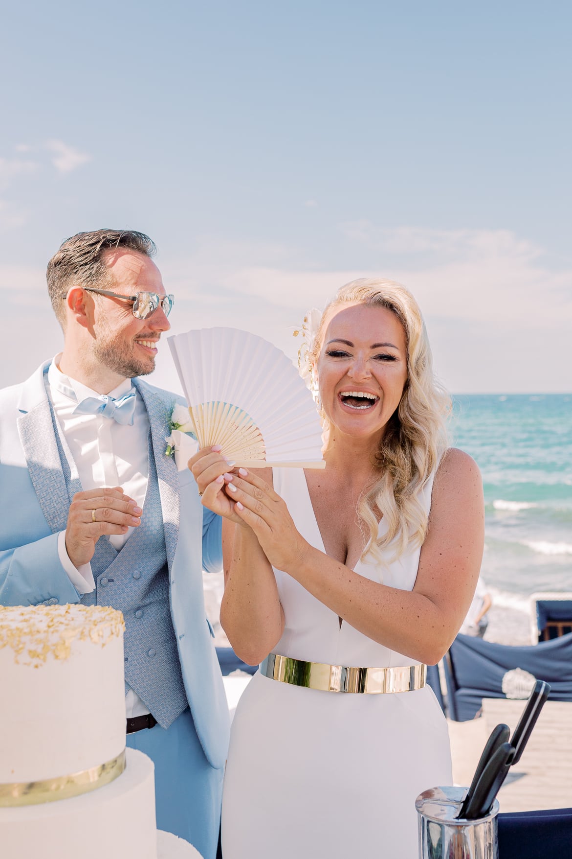 Brautpaar lacht und vor ihnen steht die Hochzeitstorte am Strand