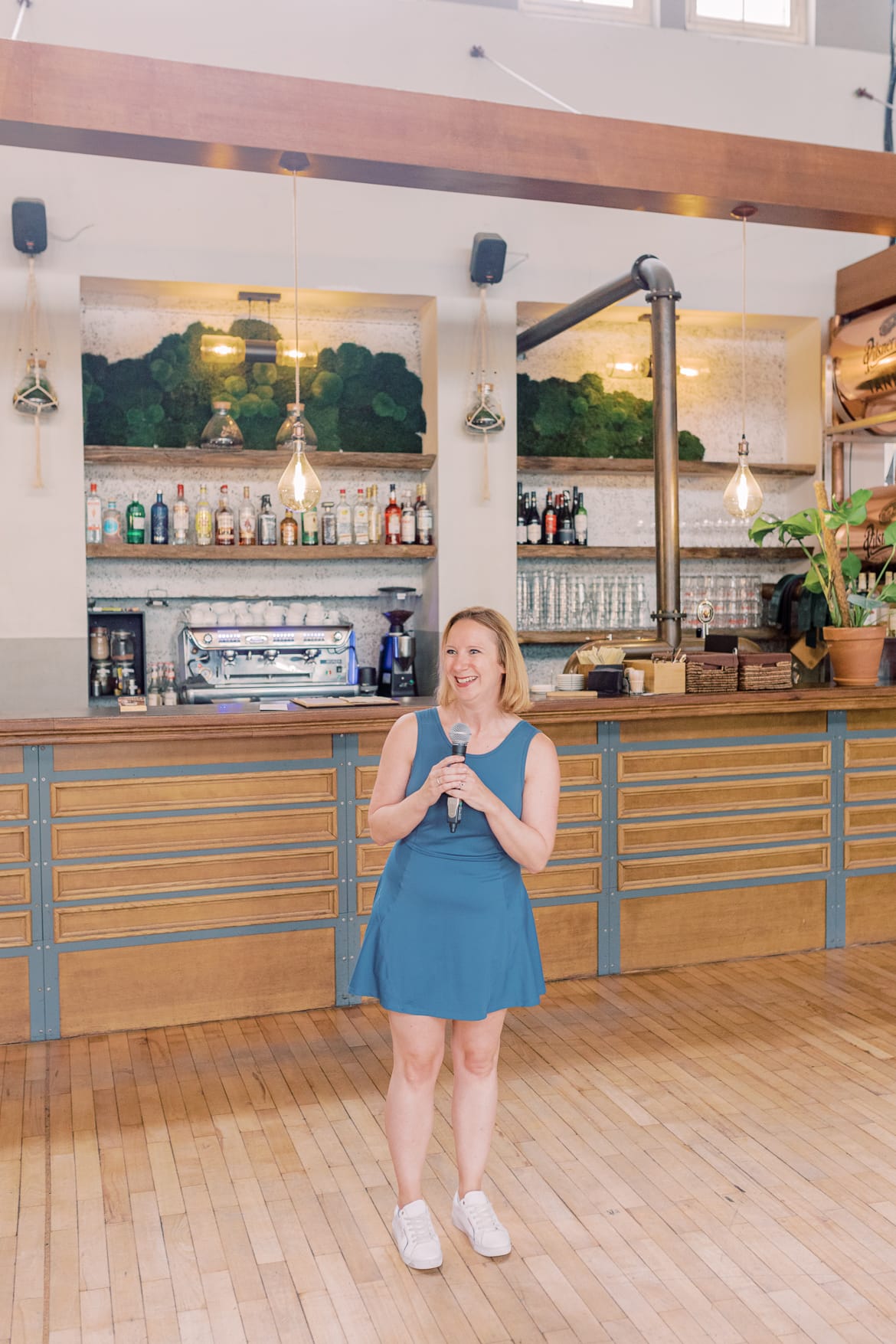 Eine blonde Frau im blauen Kleid lacht und steht vor einer Bar in einer alten Turnhalle